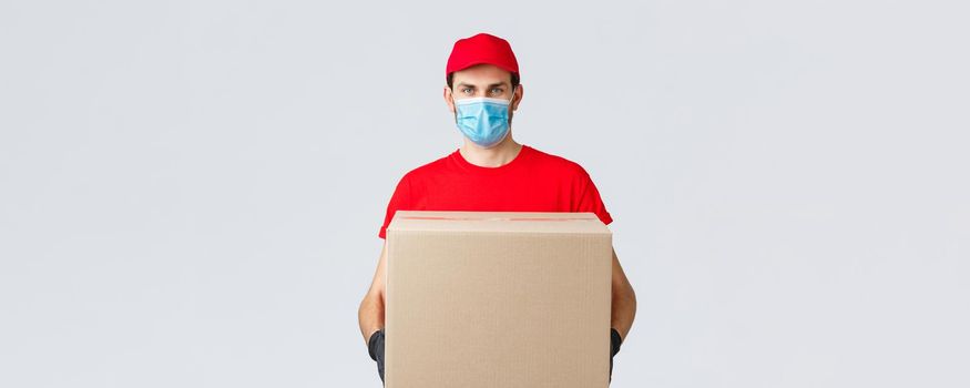 Groceries and packages delivery, covid-19, quarantine and shopping concept. Serious courier in red uniform, gloves and protective face mask, deliver package box to client house during coronavirus