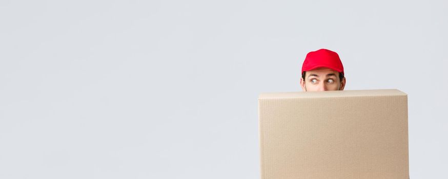 Packages and parcels delivery, covid-19 quarantine and transfer orders. Scared courier in red uniform cap, hiding behind customer order, looking left nervously, peeking at banner or advertisement
