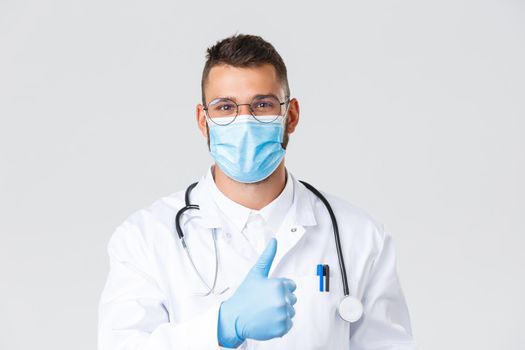 Covid-19, healthcare workers, pandemic and preventing virus concept. Handsome smiling doctor in medical mask and gloves, show thumb-up in approval, recommend clinic service or medication