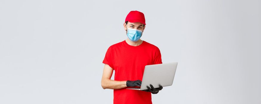 Customer support, covid-19 delivery packages, online orders processing concept. Pleasant delivery guy in red uniform, face mask and gloves, using laptop and look camera, processing order