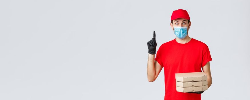 Food delivery, application, online grocery, contactless shopping and covid-19 concept. Excited delivery guy in red uniform, gloves and face mask, have suggestion, hold pizza and raise finger
