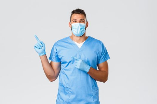 Healthcare workers, covid-19, coronavirus and preventing virus concept. Happy professional male nurse, doctor in scrubs and medical mask, gloves pointing finger left, showing banner
