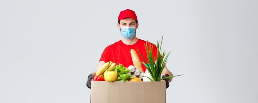 Groceries and packages delivery, covid-19, quarantine and shopping concept. Smiling courier in face mask, gloves and red uniform holding box with food, customer order, client home delivery