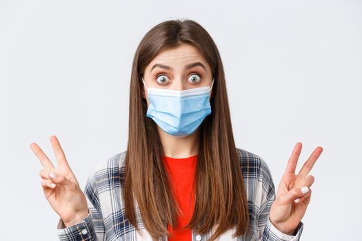 Coronavirus outbreak, leisure on quarantine, social distancing and emotions concept. Close-up of excited and thrilled cute woman in medical mask, showing peace sign or quotes, stare impressed