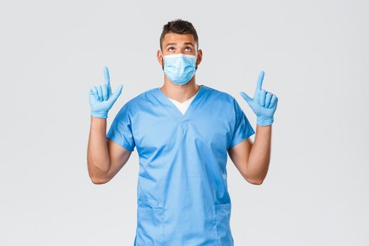 Healthcare workers, covid-19, coronavirus and preventing virus concept. Handsome hispanic male doctor, nurse in scrubs and medical mask, pointing fingers up, reading interesting news