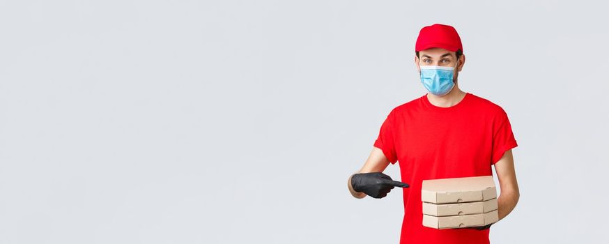 Food delivery, application, online grocery, contactless shopping and covid-19 concept. Cheerful courier in red uniform, face mask and gloves pointing finger at pizza boxes, deliver to client house