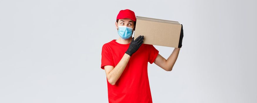 Packages and parcels delivery, covid-19 quarantine delivery, transfer orders. Curious courier in red uniform, gloves and protective face mask, deliver box to client, bring order contactless