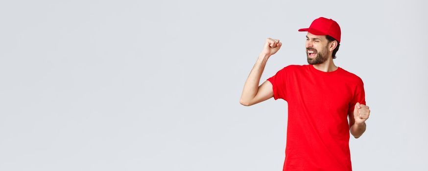Cheerful and carefree young bearded delivery guy in red uniform cap and t-shirt, singing, yelling yes and fist pump in celebration, triumphing and rejoicing over great news, grey background