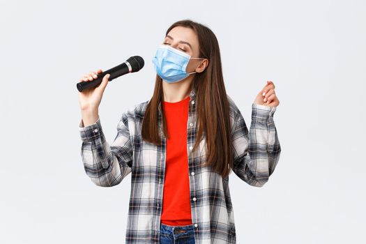 Coronavirus outbreak, leisure on quarantine, social distancing and emotions concept. Carefree young girl in medical mask, singing with microphone, close eyes, enjoying karaoke