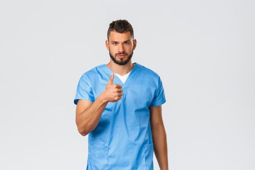 Healthcare workers, medicine, covid-19 and pandemic self-quarantine concept. Determined handsome doctor, male nurse in blue scrubs, thumb-up in support, save patients, assure quality service