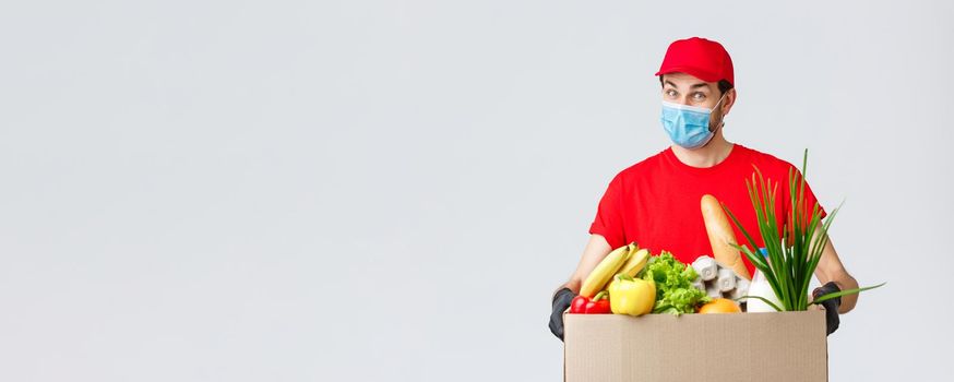 Groceries and packages delivery, covid-19, quarantine and shopping concept. Friendly courier in face mask and gloves, red uniform bring food box to customer ordered online, contactless deliver