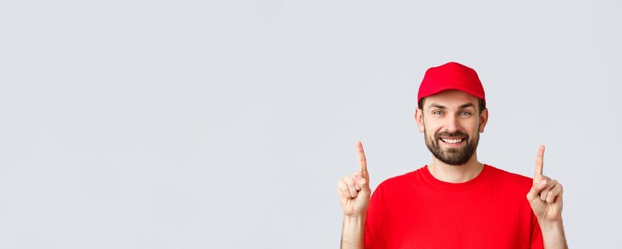 Online shopping, delivery during quarantine and takeaway concept. Cheerful bearded smiling courier in red uniform cap and t-shirt, invite take look at promo, pointing fingers up, grey background