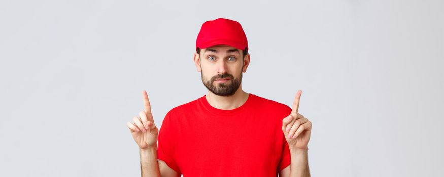 Online shopping, delivery during quarantine and takeaway concept. Indecisive and unsure courier in red uniform cap, t-shirt, smirk and pointing fingers up uncertain, dont know, cant decide