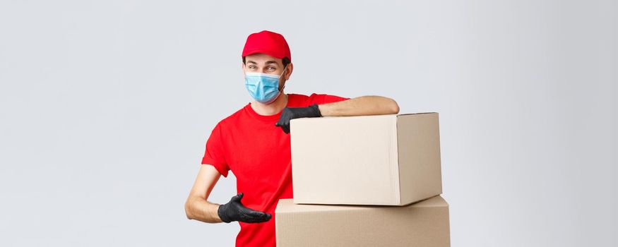 Packages and parcels delivery, covid-19 quarantine and transfer orders. Smiling courier in red uniform, gloves and medical face mask, introduce boxes to transfer your order, recommend service