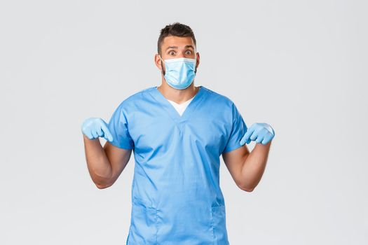 Healthcare workers, covid-19, coronavirus and preventing virus concept. Confused and surprised handsome male doctor, nurse in medical mask, gloves and scrubs pointing fingers down at advertisement