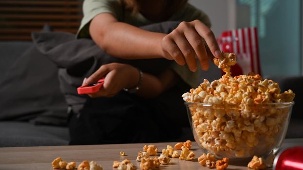 Shot of woman hand grabbing popcorn from a wooden bowl and watching TV at home. Leisure activity concept