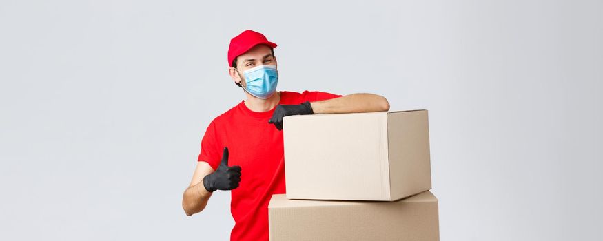 Packages and parcels delivery, covid-19 quarantine and transfer orders. Confident courier in red uniform, gloves and medical mask, encourage call service, show thumb-up lean on boxes