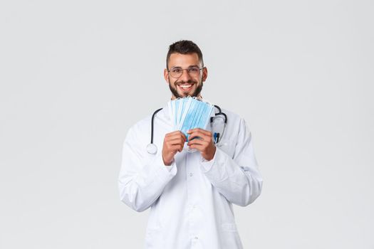 Healthcare workers, medical insurance, pandemic and covid-19 concept. Happy good-looking doctor in glasses and scrubs, showing medical masks and smiling, recommend protect yourself during corona