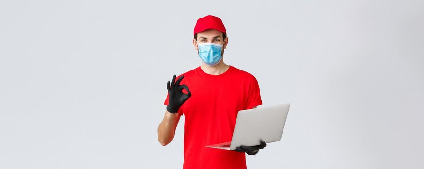Customer support, covid-19 delivery packages, online orders processing concept. Smiling courier in face mask and gloves guarantee safety of parcel, processing order, show okay sign, hold laptop