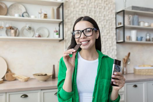 Young beautiful brunette woman in glasses and green shirt at home in kitchen, happy and smiling looking at camera eating dark chocolate