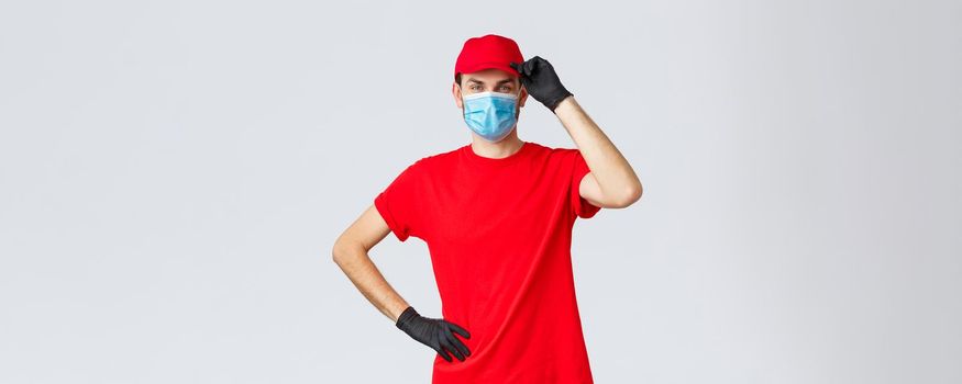 Covid-19, self-quarantine, online shopping and shipping concept. Delivery guy red uniform, touching cap as saluting customer, working in coronavirus outbreak, wear medical mask and protective gloves