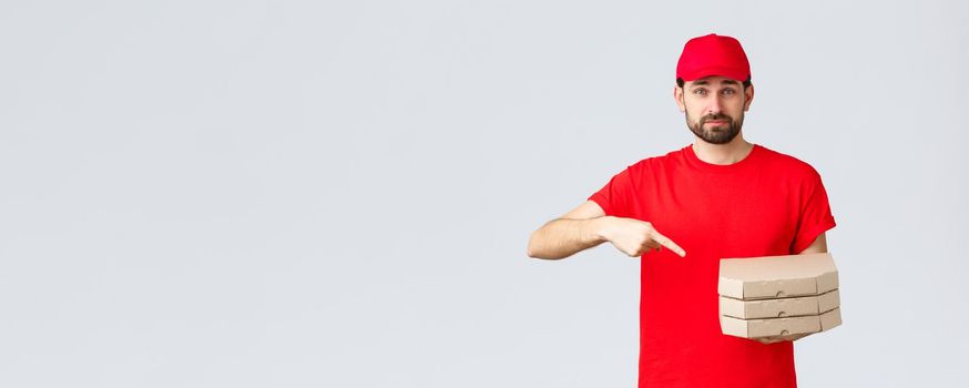 Food delivery, quarantine, stay home and order online concept. Confident friendly courier in red uniform cap and t-shirt, employee bring order pizza, pointing finger at boxes, grey background