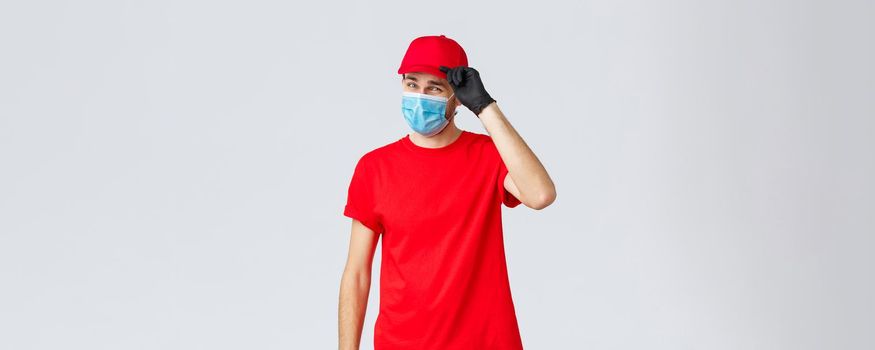 Covid-19, self-quarantine, online shopping and shipping concept. Sassy delivery man in red uniform and mask with gloves, saluting client, deliver packages and orders during coronavirus, food orders