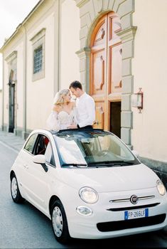 Milan, Italy - 07.04.21: Bride and groom stand in the hatch of the car Fiat 500 near the building