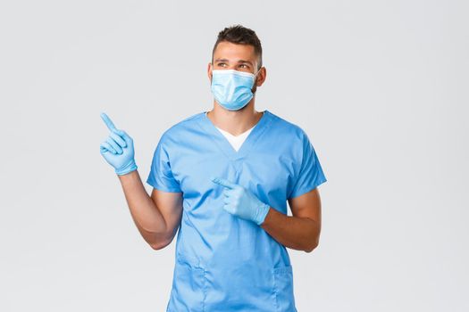 Healthcare workers, covid-19, coronavirus and preventing virus concept. Young happy doctor, handsome male nurse in scrubs and medical mask, pointing looking upper left corner with pleased smile