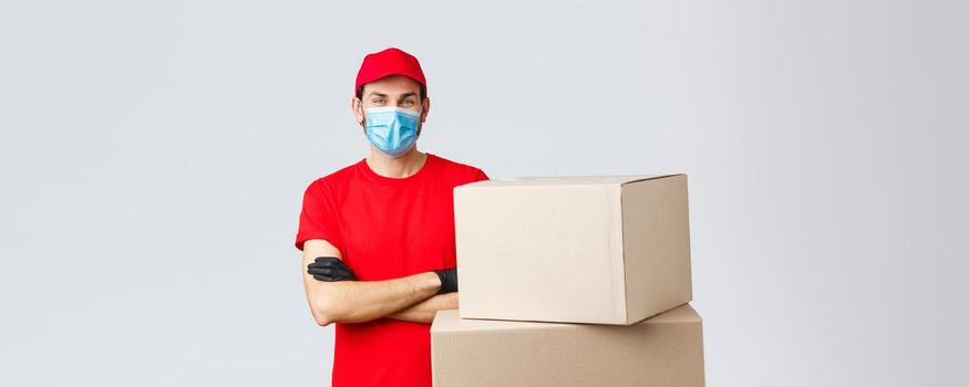 Packages and parcels delivery, covid-19 quarantine and transfer orders. Confident young courier in red uniform, gloves and medical mask, cross arms as standing boxes, grey background