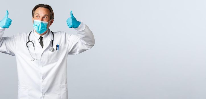 Covid-19, preventing virus, healthcare workers and vaccination concept. Cheerful pleased doctor in medical mask and gloves show thumbs-up and looking left satisfied, white background