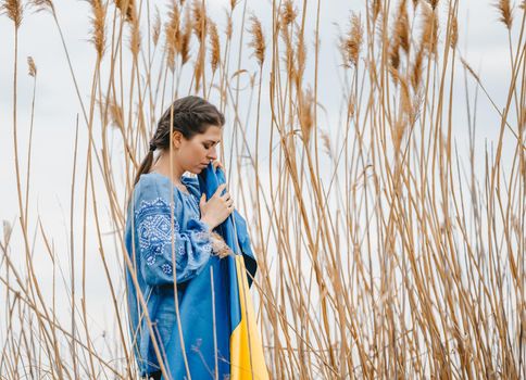 Sorrowful ukrainian woman with national flag on natural reeds background. Lady in blue embroidery vyshyvanka blouse. Ukraine, independence, freedom, patriot symbol, victory in war.
