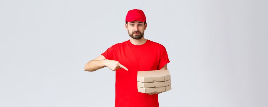 Food delivery, quarantine, stay home and order online concept. Confident friendly courier in red uniform cap and t-shirt, employee bring order pizza, pointing finger at boxes, grey background