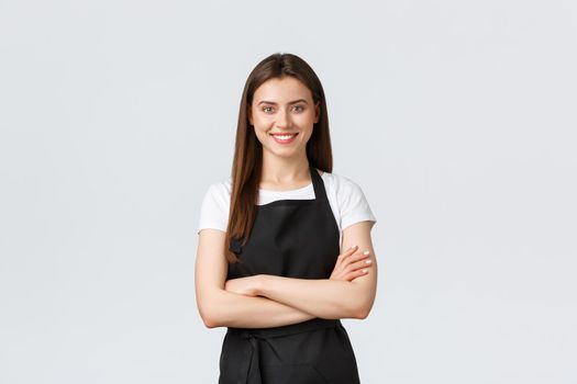 Cheerful smiling female barista in black apron cross arms chest, looking ready and confident. Young girl employee open coffee shop, greeting customers. Saleswoman running store
