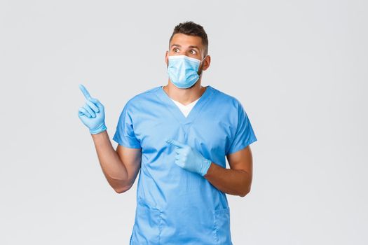 Healthcare workers, covid-19, coronavirus and preventing virus concept. Shocked and startled handsome doctor, male nurse in scrubs and medical mask, pointing fingers upper left corner and read banner