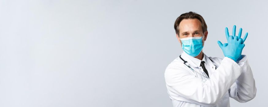 Covid-19, preventing virus, healthcare workers and vaccination concept. Talented smiling doctor in medical mask and gloves, hold hands together and looking camera optimistic.