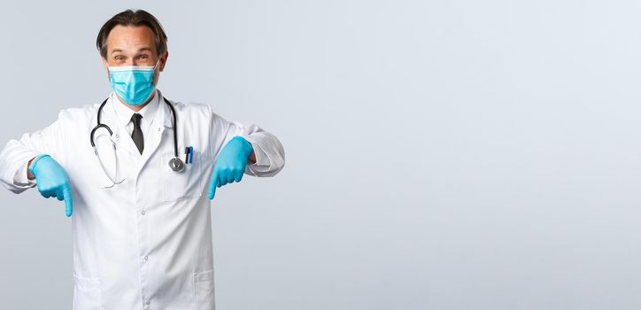 Covid-19, preventing virus, healthcare workers and vaccination concept. Enthusiastic handsome male doctor in medial mask and gloves, pointing fingers down at advertisement, white background.
