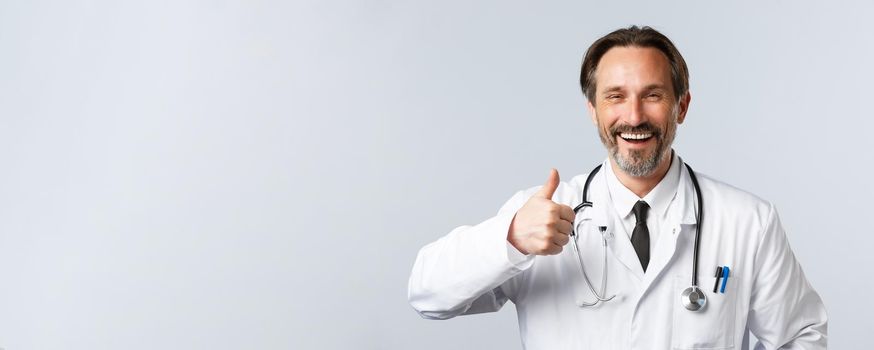 Covid-19, preventing virus, healthcare workers and vaccination concept. Cheerful middle-aged male doctor in white coat laughing happy and thumb-up in approval, recommend product or services