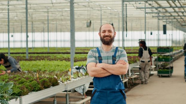 Portrait of smiling man posing with arms crossed standing in greenhouse with diverse workers pushing crates
