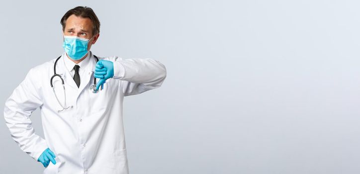 Covid-19, preventing virus, healthcare workers and vaccination concept. Skeptical and disappointed doctor in medical mask, gloves show thumb-down displeased, look top left sad.