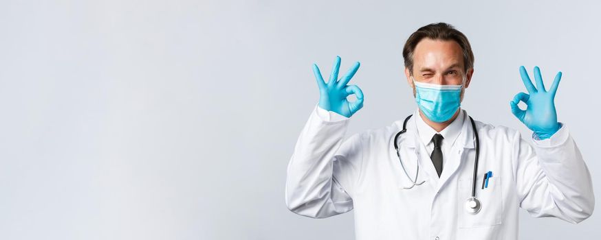 Covid-19, preventing virus, healthcare workers and vaccination concept. Friendly smiling doctor in medical mask and gloves, wink cheeky and show okay gesture, guarantee or recommend service