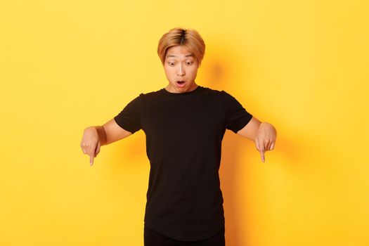 Portrait of surpirsed asian guy gasping amazed, pointing fingers and looking down at banner, standing yellow background