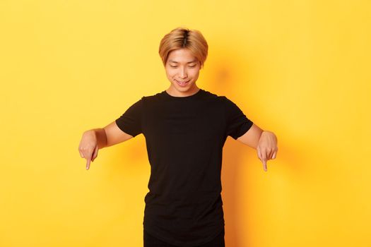 Portrait of curious happy blond asian guy, looking and pointing fingers down with pleased smile, yellow background