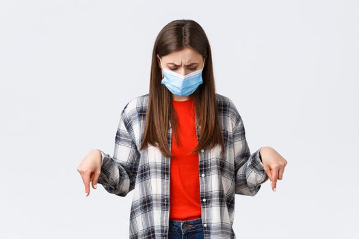 Coronavirus outbreak, leisure on quarantine, social distancing and emotions concept. Confused and unsure young woman in medical mask stare puzzled at something down, point bottom promo