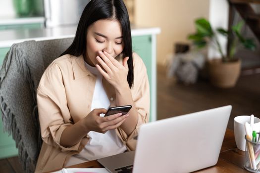Happy asian girl sitting with laptop at home, looking at mobile phone and laughing, has workplace at house