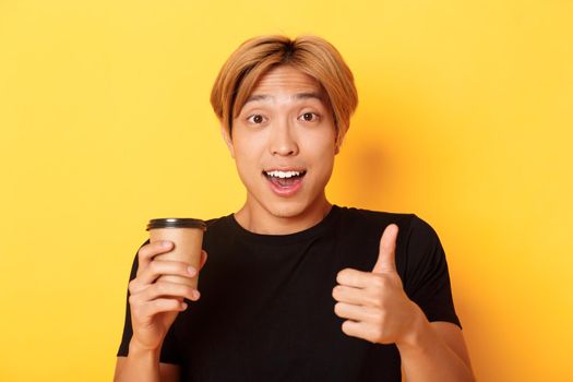 Close-up of surprised asian handsome guy recommend cafe, holding cup of coffee and showing thumbs-up in approval, smiling pleased over yellow background