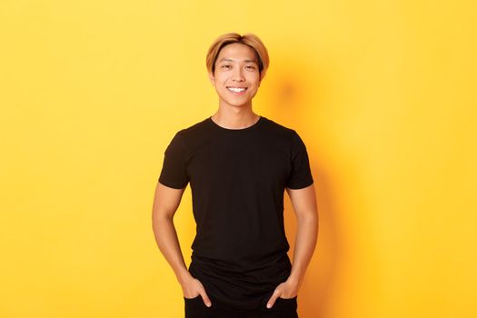 Attractive smiling asian man in black t-shirt, standing pleased, yellow background