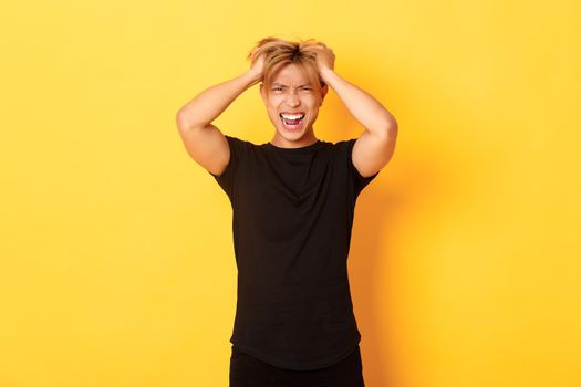 Portrait of pissed-off angry asian man tossing haircut and yelling furious, standing over yellow background