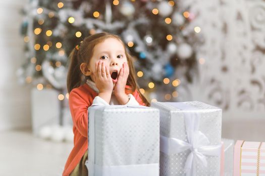 Cute little girl surprised by the abundance of Christmas gifts by a Christmas tree in cozy room