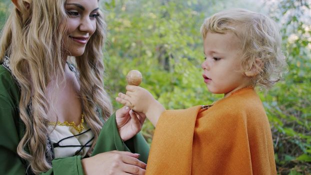 Fairy elf cosplay woman treats little baby boy hobbit with candy in green forest. Halloween concept, fairytale characters, kids.